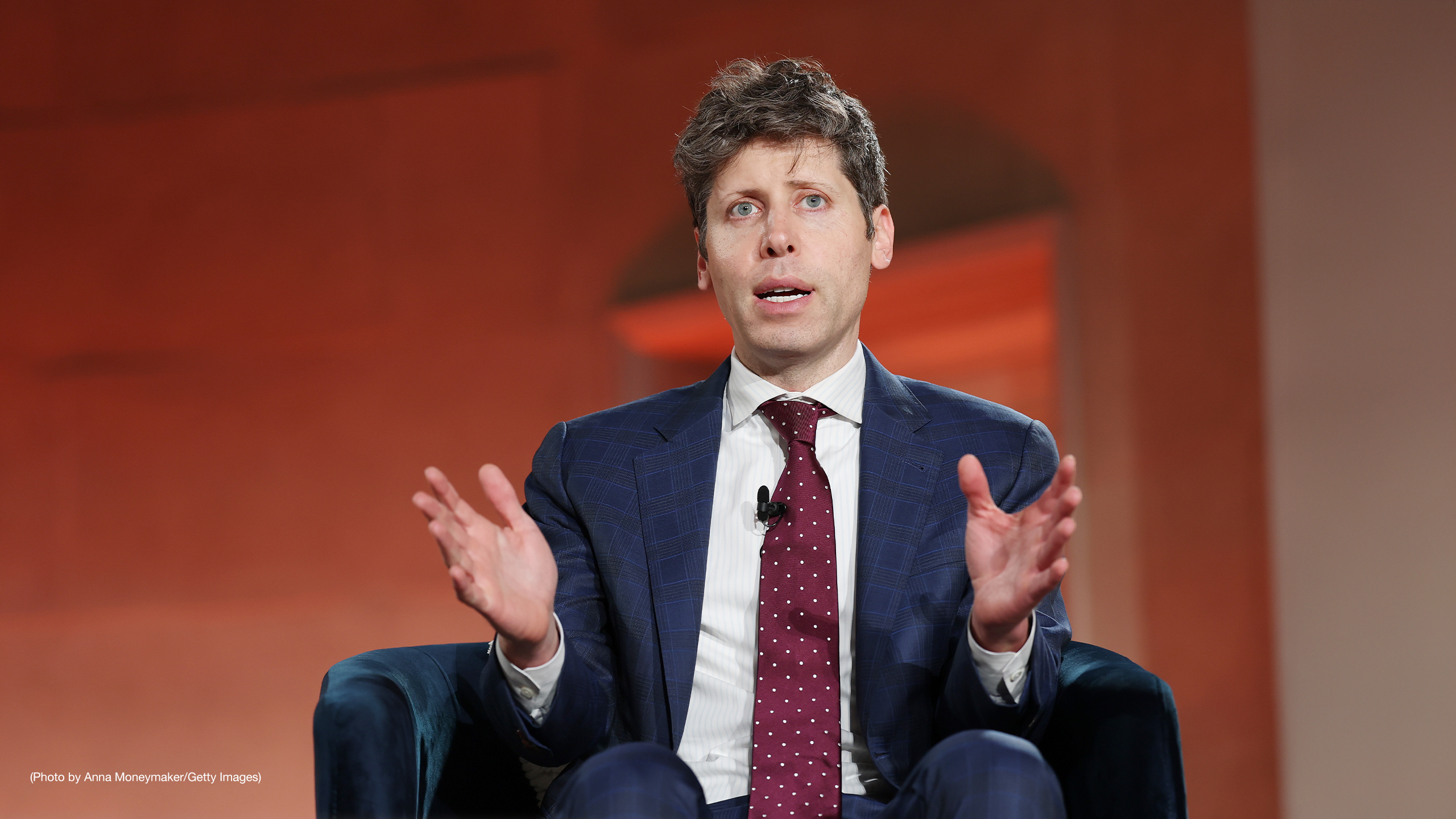 OpenAI Sam Altman speaking in a blue suit, red tie, with his hands up, in front of a dark red background